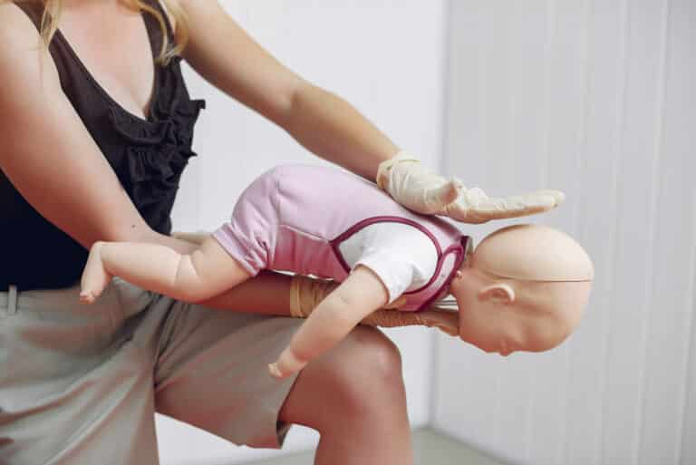 Rückenschläge bei einem Säugling: Ersthelferin übt an einer Babypuppe in Bauchlage auf dem Unterarm