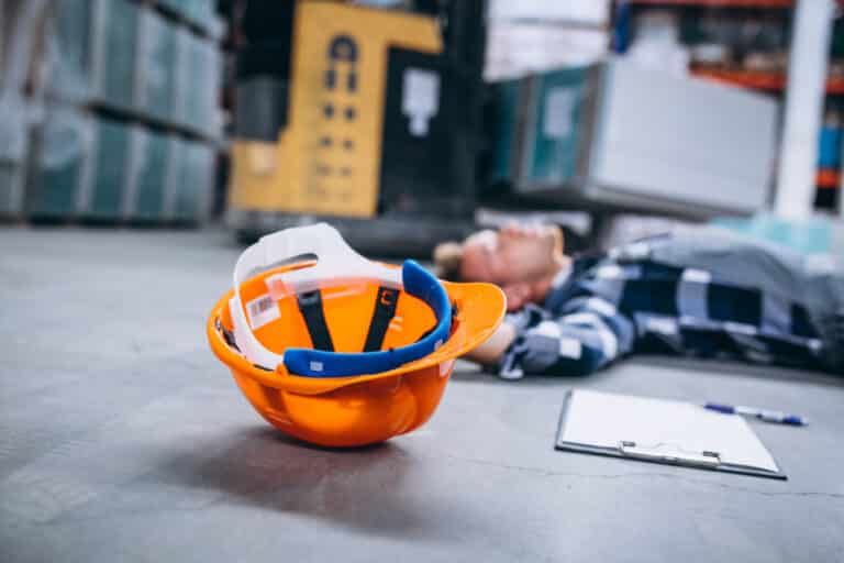 Arbeitsunfall im Lager: Beschäftigter liegt am Boden, im Vordergrund sein Schutzhelm