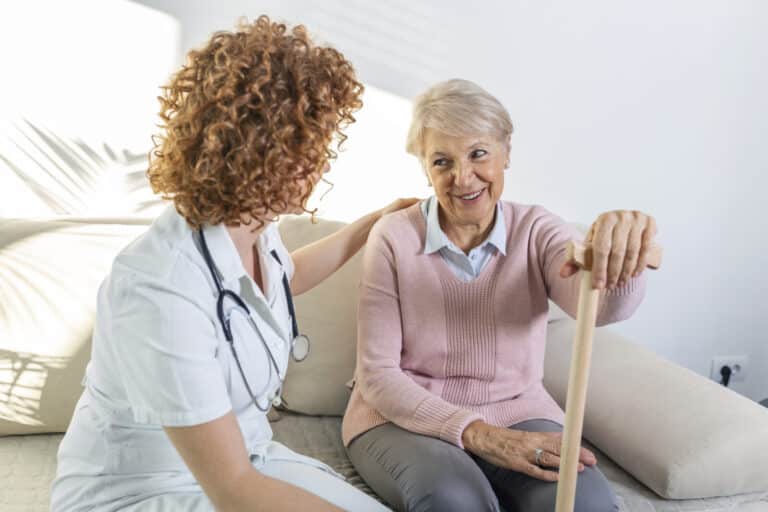 Pflegekraft im Gespräch mit einer lächelnden Seniorin mit Gehstock